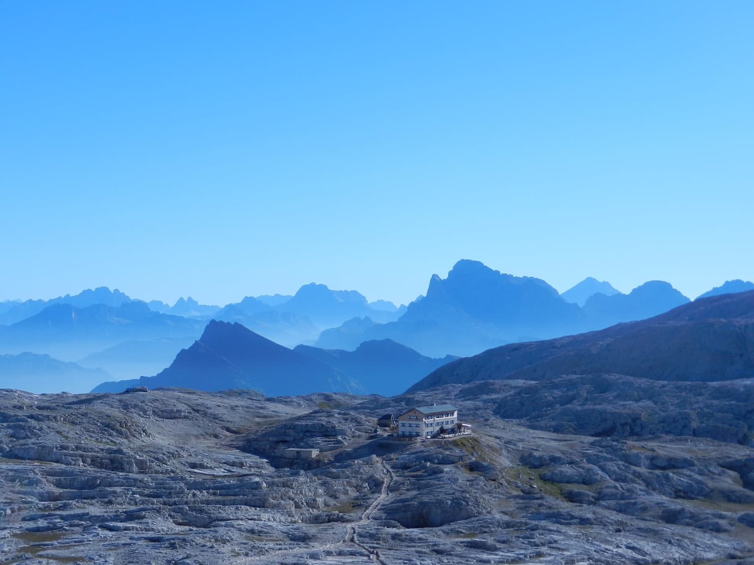 Rifugio Rosetta standing alone on the bare limestone of the Pale di San Martino plateau, a footpath crossing the gray rock toward it and hazy blue ridges beyond