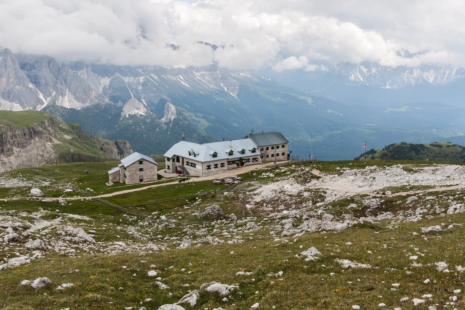 Rifugio Bolzano, a long gray stone hut with a smaller annex, on the rock-strewn grass of the Sciliar plateau, snow-streaked ranges under a heavy cloud bank behind