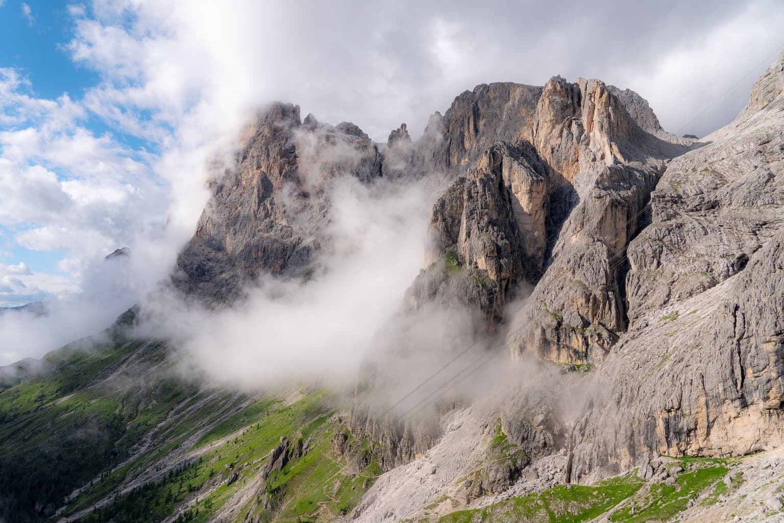 Thick cloud billows up and over a jagged Dolomite ridge, swallowing the peak's upper towers, while a patch of clear blue sky and green alpine slope remain visible to the side.