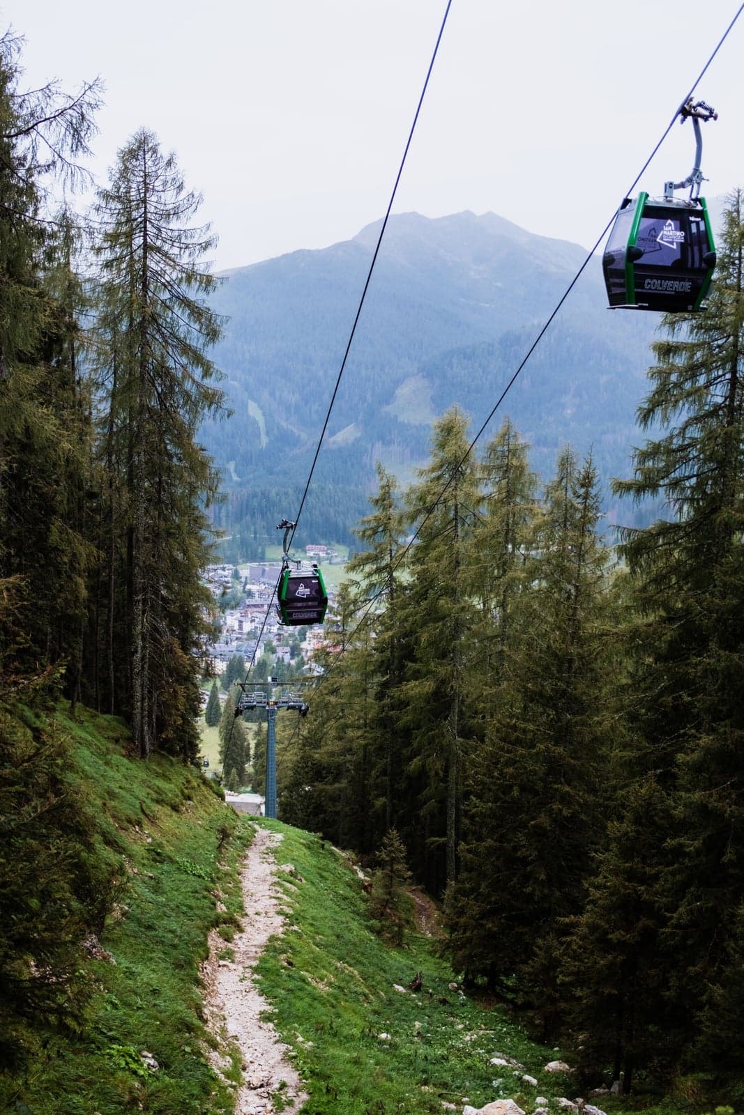 A cable-car cabin descends a forested line above a hiking trail, with the roofs of a valley village and a mountain ridge visible below.