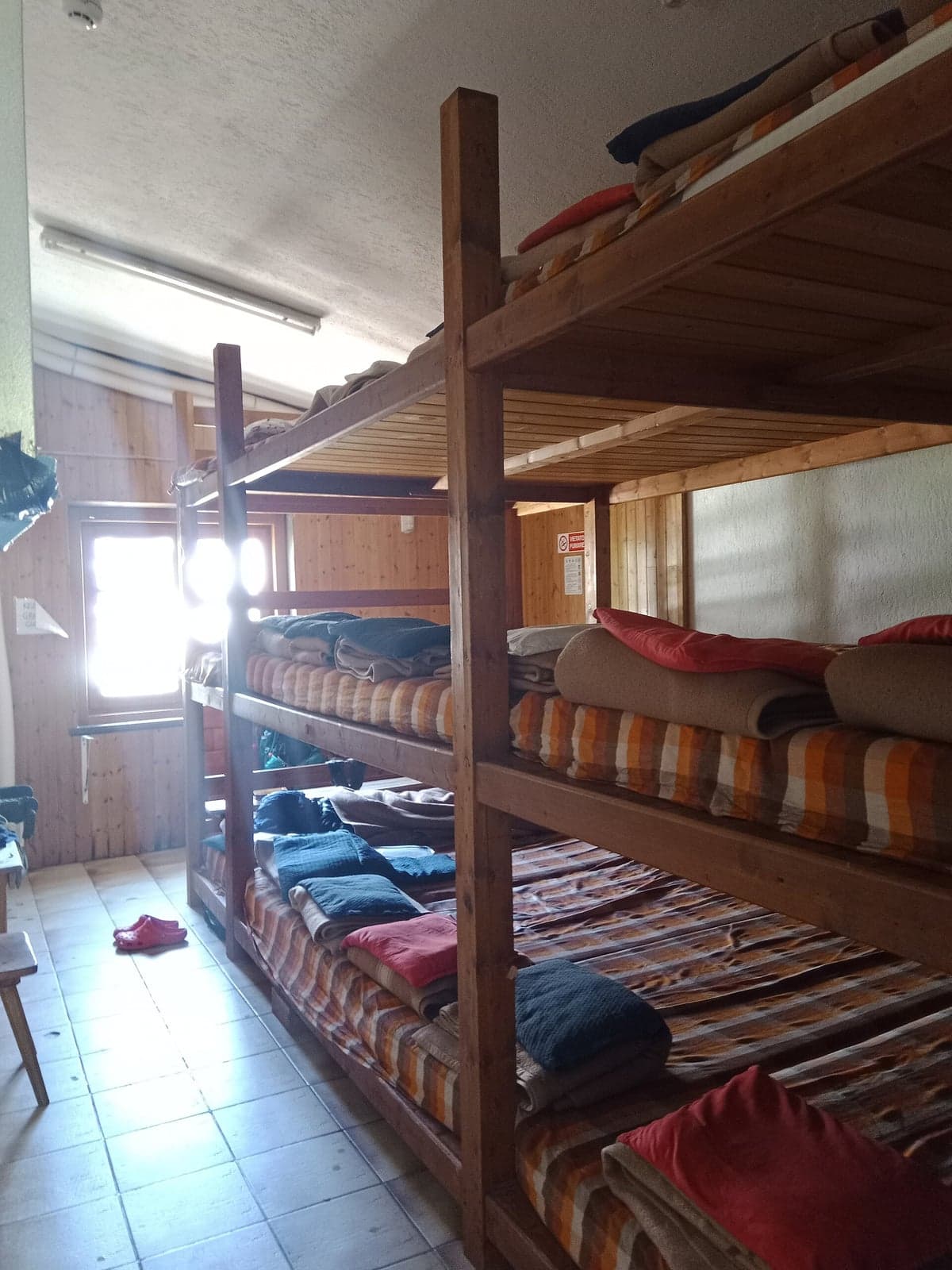 Inside a mountain-hut dormitory: stacked wooden bunk beds with striped mattresses, wool blankets folded at the foot of each bed, pillows stacked at the head, a pair of slippers left on the tiled floor, morning light through a small window.