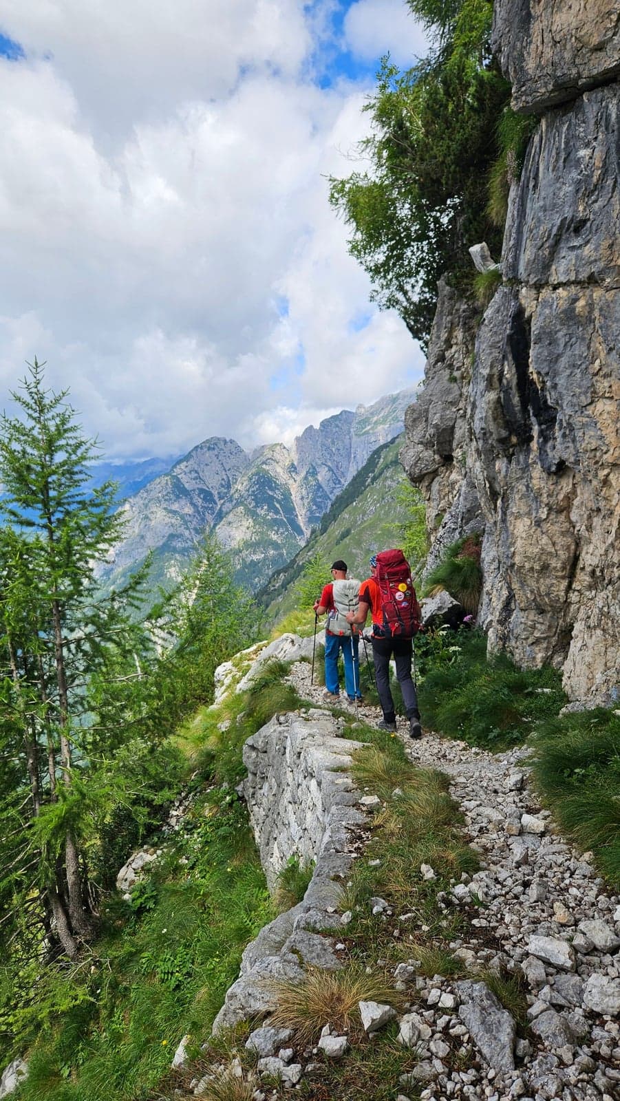 Two hikers with backpacks and trekking poles walk single file along a narrow trail ledge reinforced by a low stone retaining wall, cut into a steep rock face, with jagged gray peaks visible across the valley.