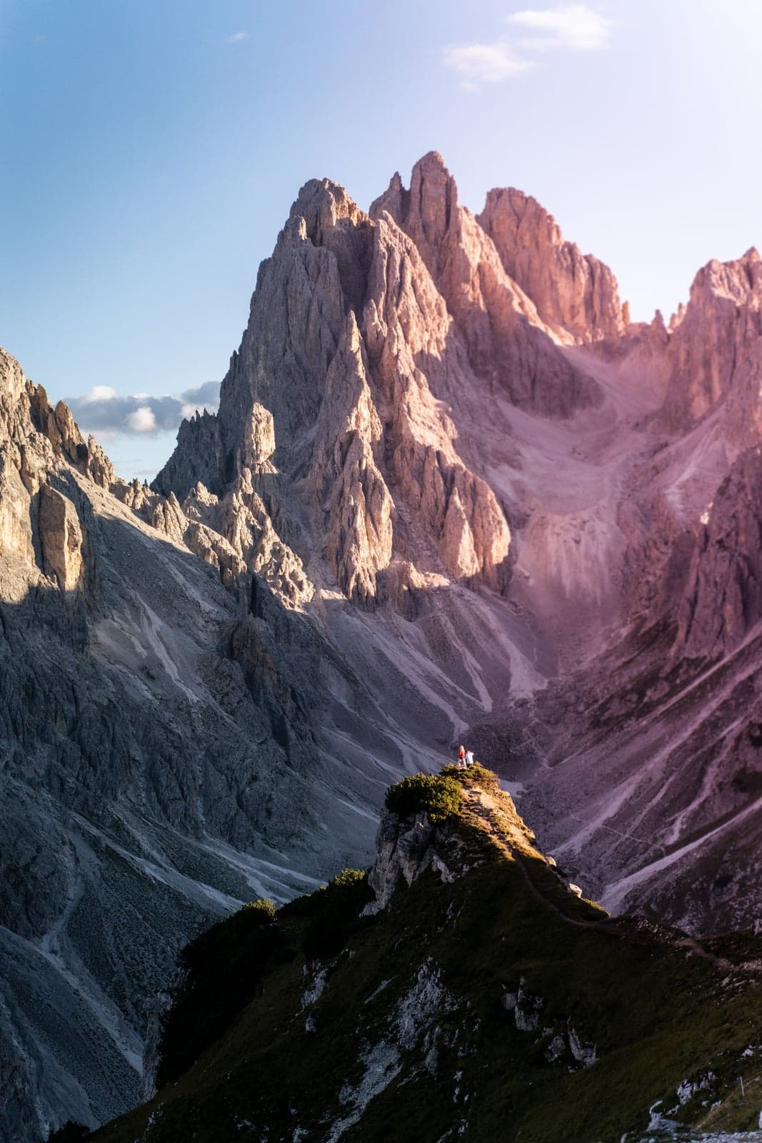 Two hikers stand on a narrow rocky outcrop lit pink by low sun, a footpath visible leading up to the vantage point, a row of sharp Dolomite towers rising behind them and a scree valley in shadow below.