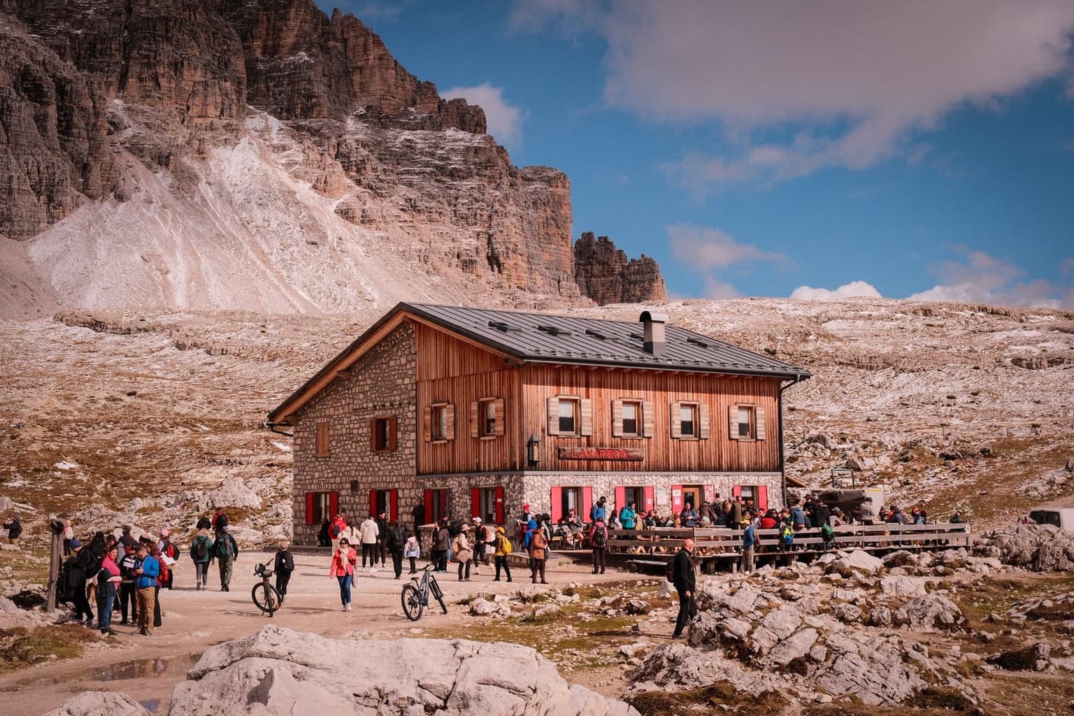 A crowded stone-and-timber rifugio, red shutters open, its terrace and yard packed with hikers and a few parked bicycles near the entrance, sheer rock cliffs rising behind.