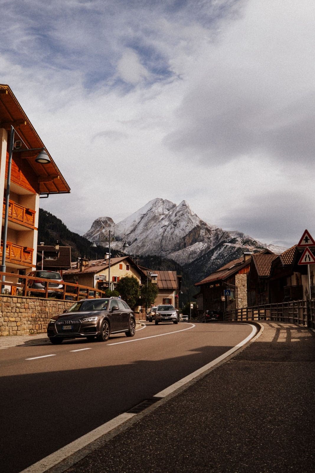 A gray SUV drives down a South Tyrolean village street lined with wood-balconied chalet buildings, a snow-dusted dolomite peak rising directly behind the rooftops.