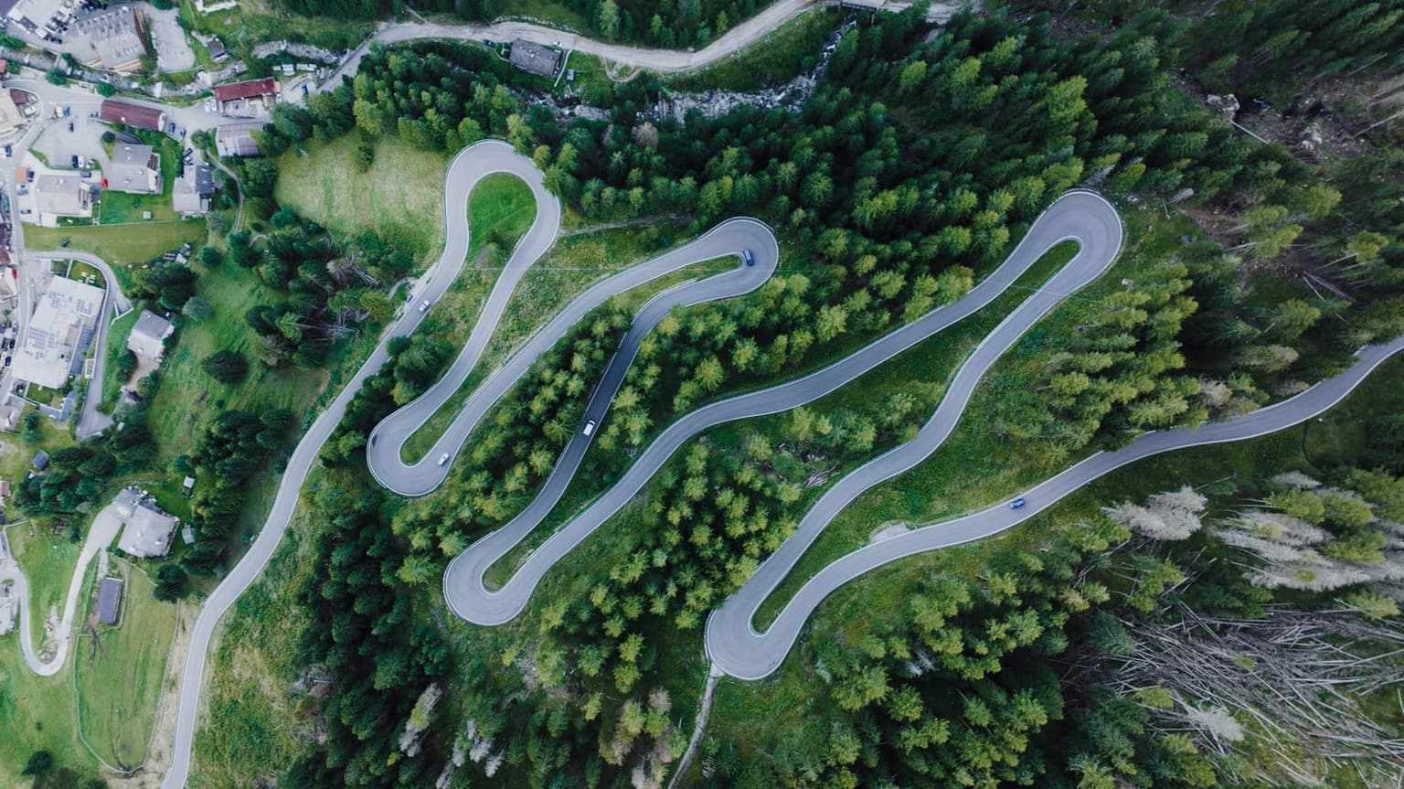 Aerial view of a paved mountain road with three tight hairpin switchbacks through pine forest on a Dolomite pass, a small village and open valley visible at the bottom of the frame, a few cars on the bends.