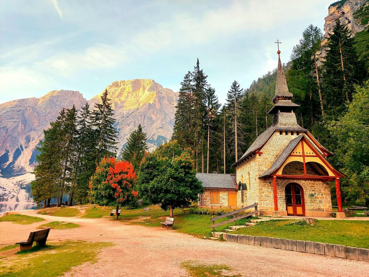 The small stone chapel beside Lago di Braies, a shingled spire and red-timbered porch, ringed by pines and autumn foliage, a Dolomite peak catching golden morning light behind