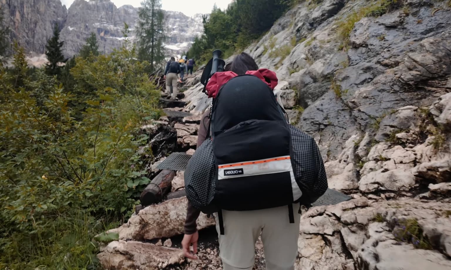 A hiker crosses a rock-and-plank trail section as a line of several more hikers climbs ahead toward misty peaks.