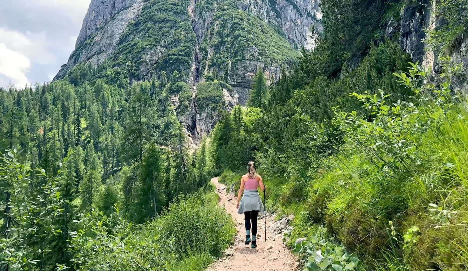 A hiker walks a shaded forest trail lined with spruce and larch, a gray Dolomite wall rising beyond the trees.