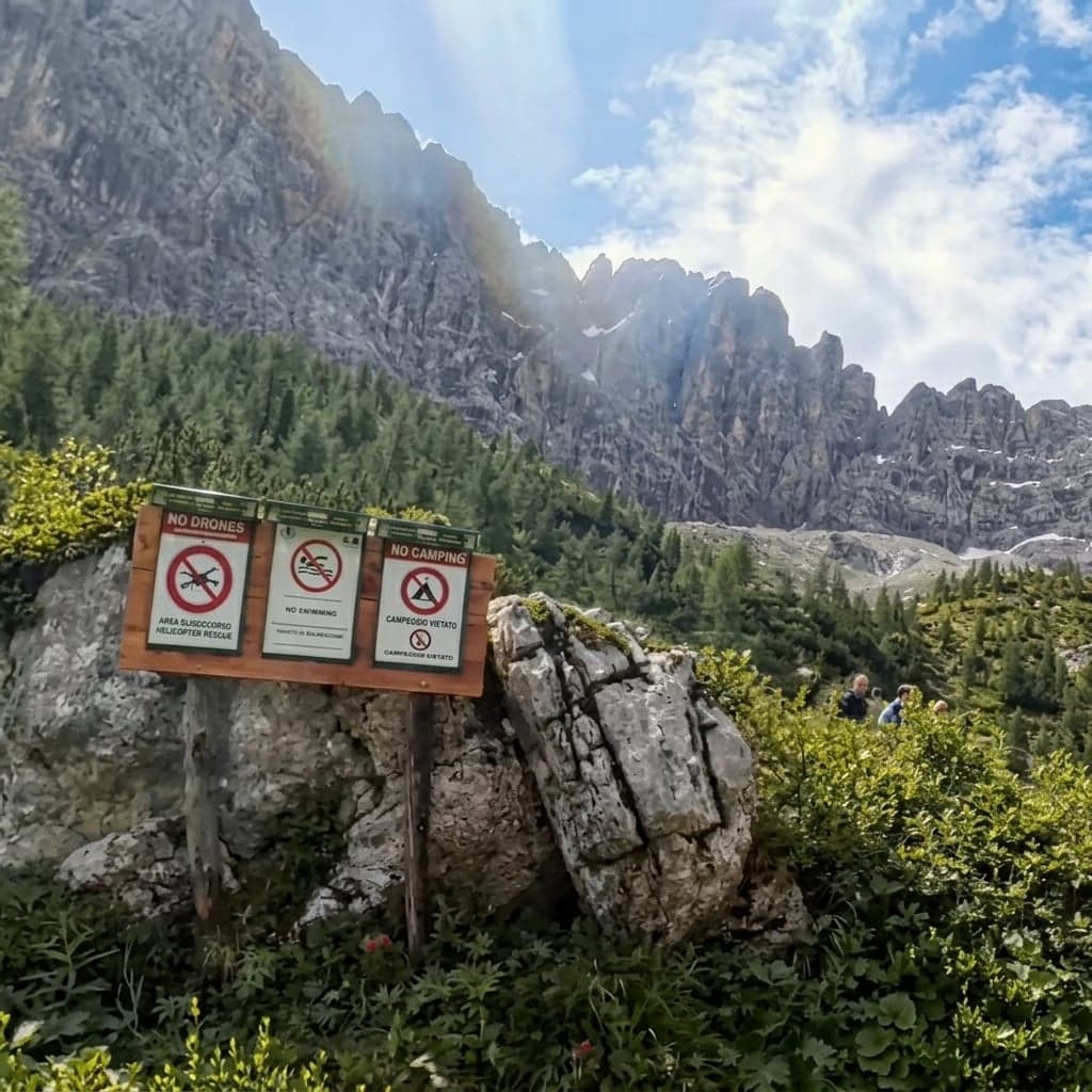 A wooden sign at the shore prohibits drones, swimming, and camping, with Dolomite peaks behind.
