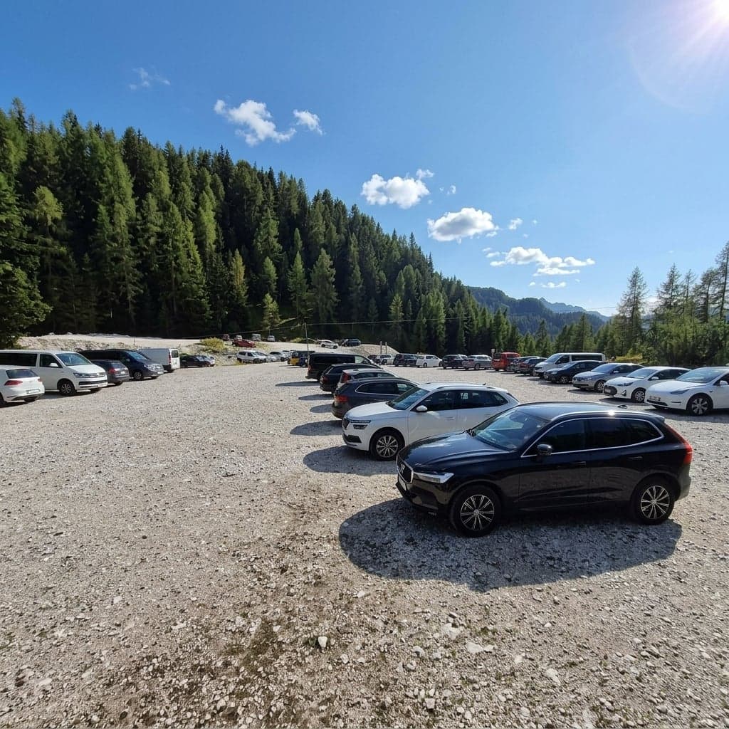 Rows of parked cars fill the gravel lot at Passo Tre Croci, pine forest and distant mountains behind.