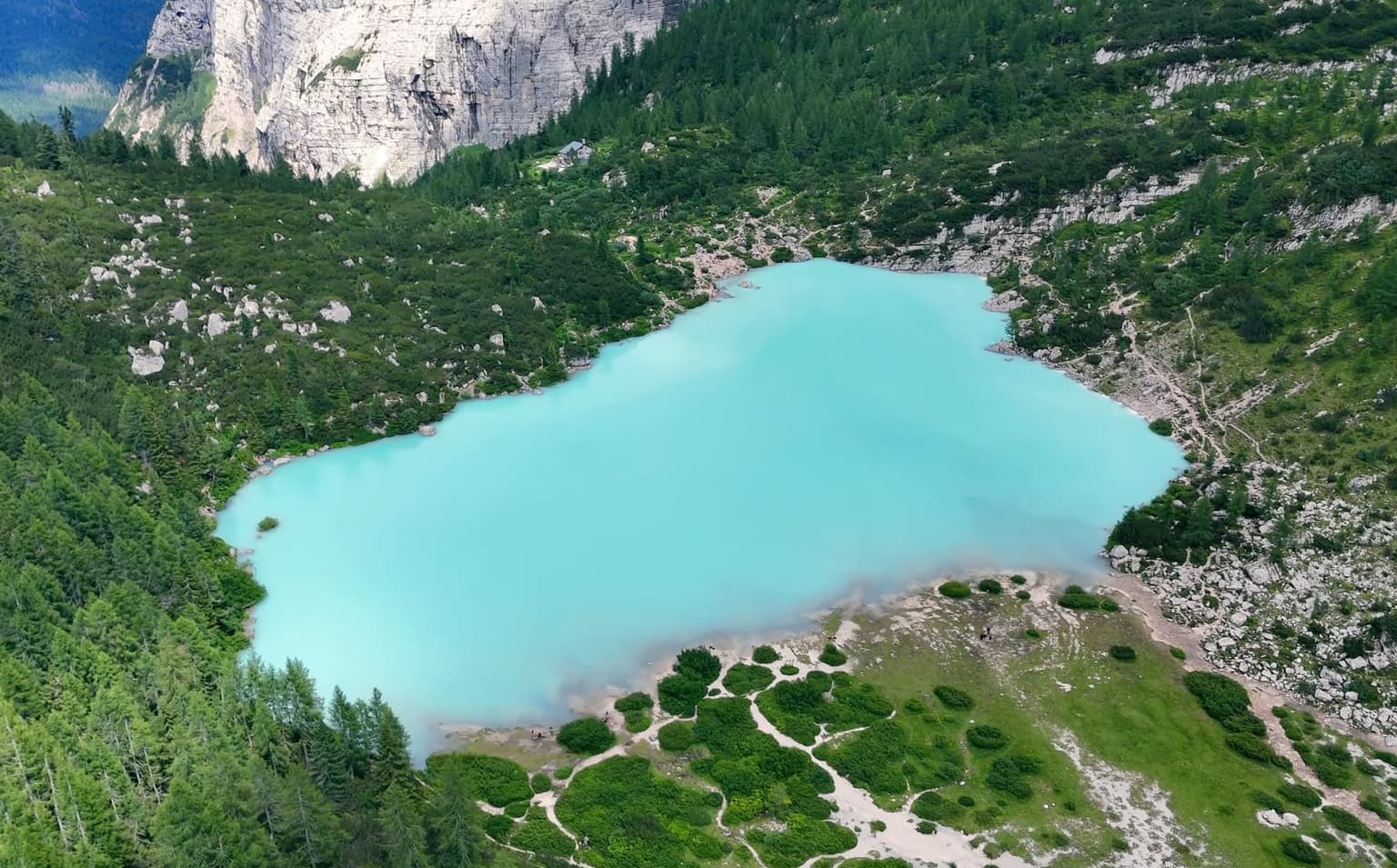 An aerial view of Lago di Sorapis shows the turquoise water fading to pale, partly exposed shoreline, forest and a rock wall around it.