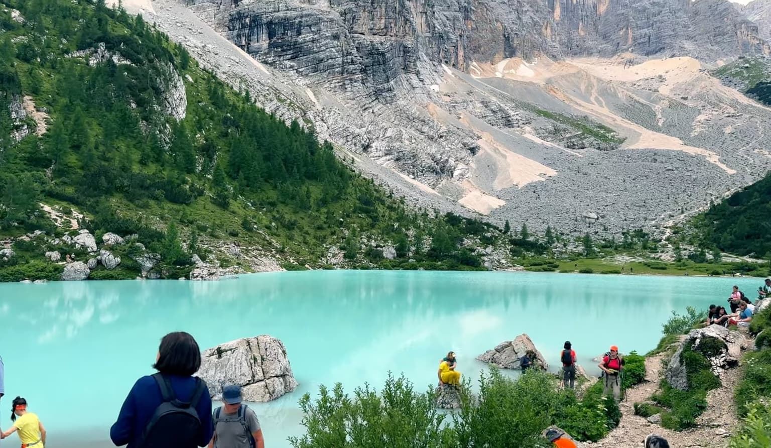 Milky-turquoise water laps pale rock at the edge of Lago di Sorapis, hikers gathered along the near shore.
