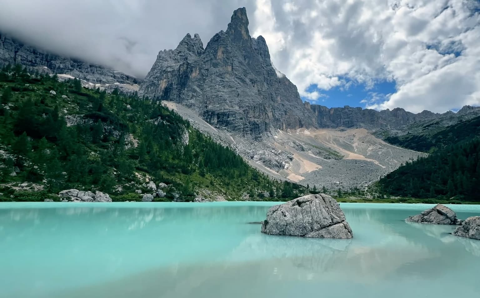 Wide view of Lago di Sorapis's milky-turquoise water, boulders breaking the surface, and the Sorapis massif's limestone spire and scree fan behind it.