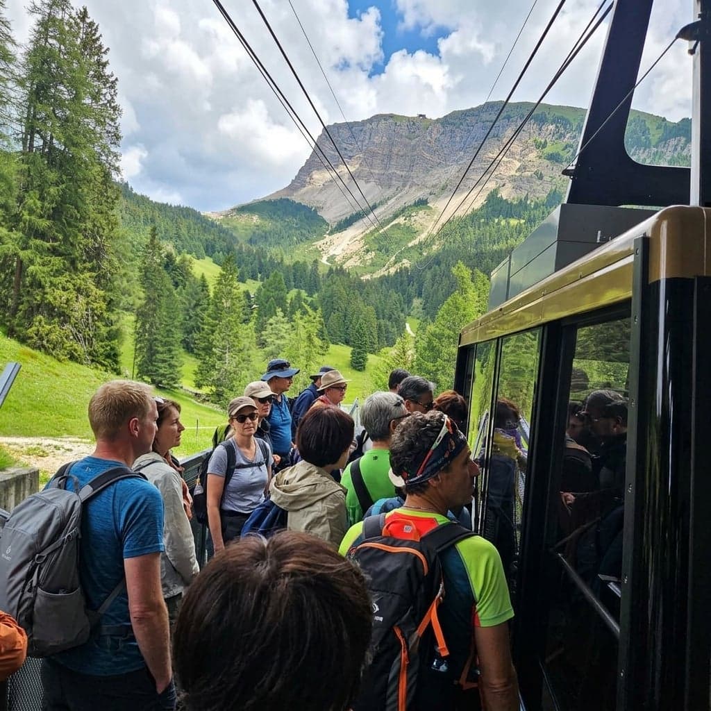 A gold-and-black cable car cabin at a mid-station on the Furnes - Seceda lift line, with a line of hikers and backpackers waiting to board; a pale rocky Dolomite peak and forested slopes are visible through the cabin's glass front.