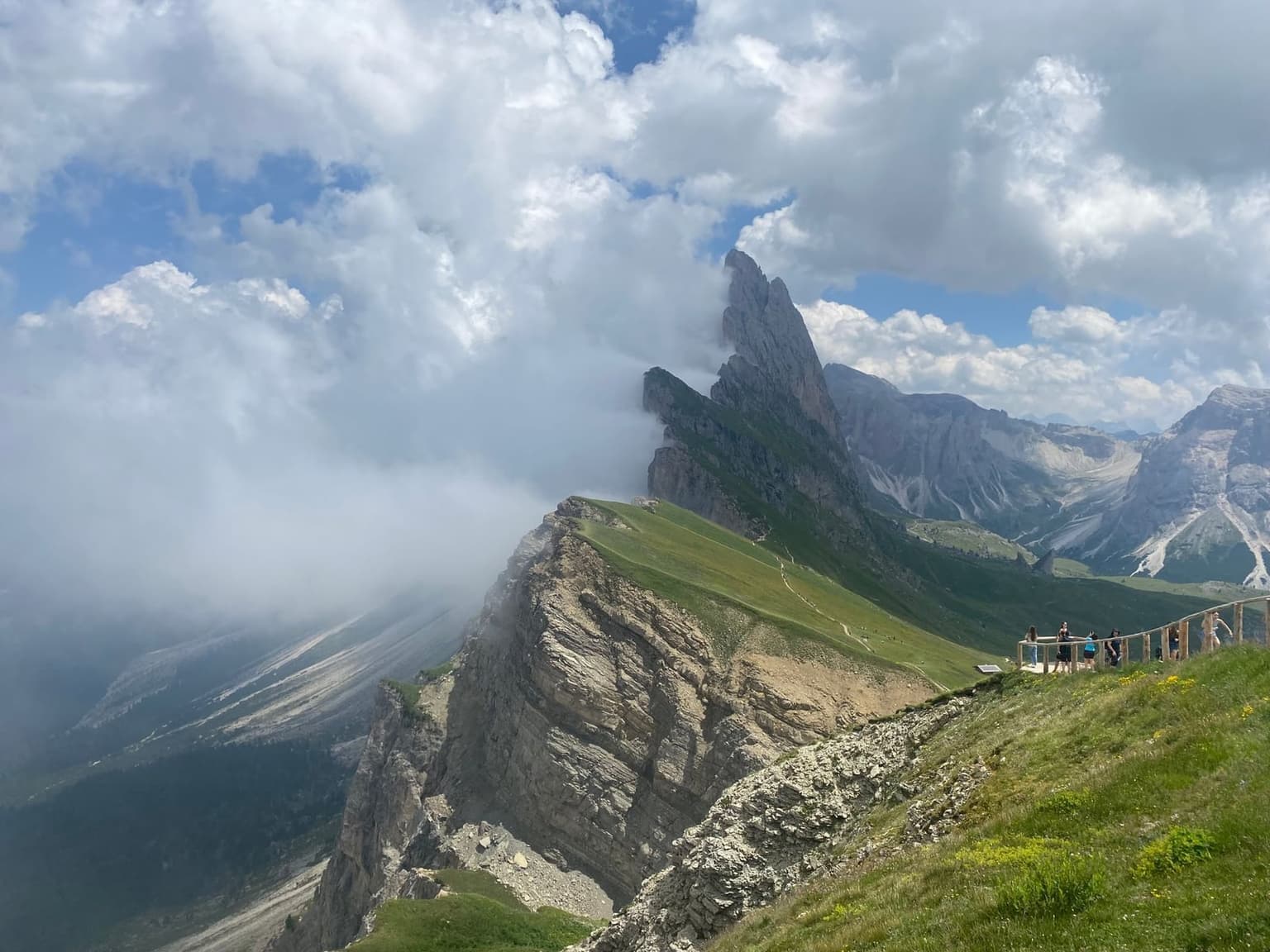 A jagged limestone spire of the Odle group breaks through a bank of cloud that has swallowed the ridge below, a second peak still clear against blue sky and hikers standing at a wooden viewing platform in the foreground.