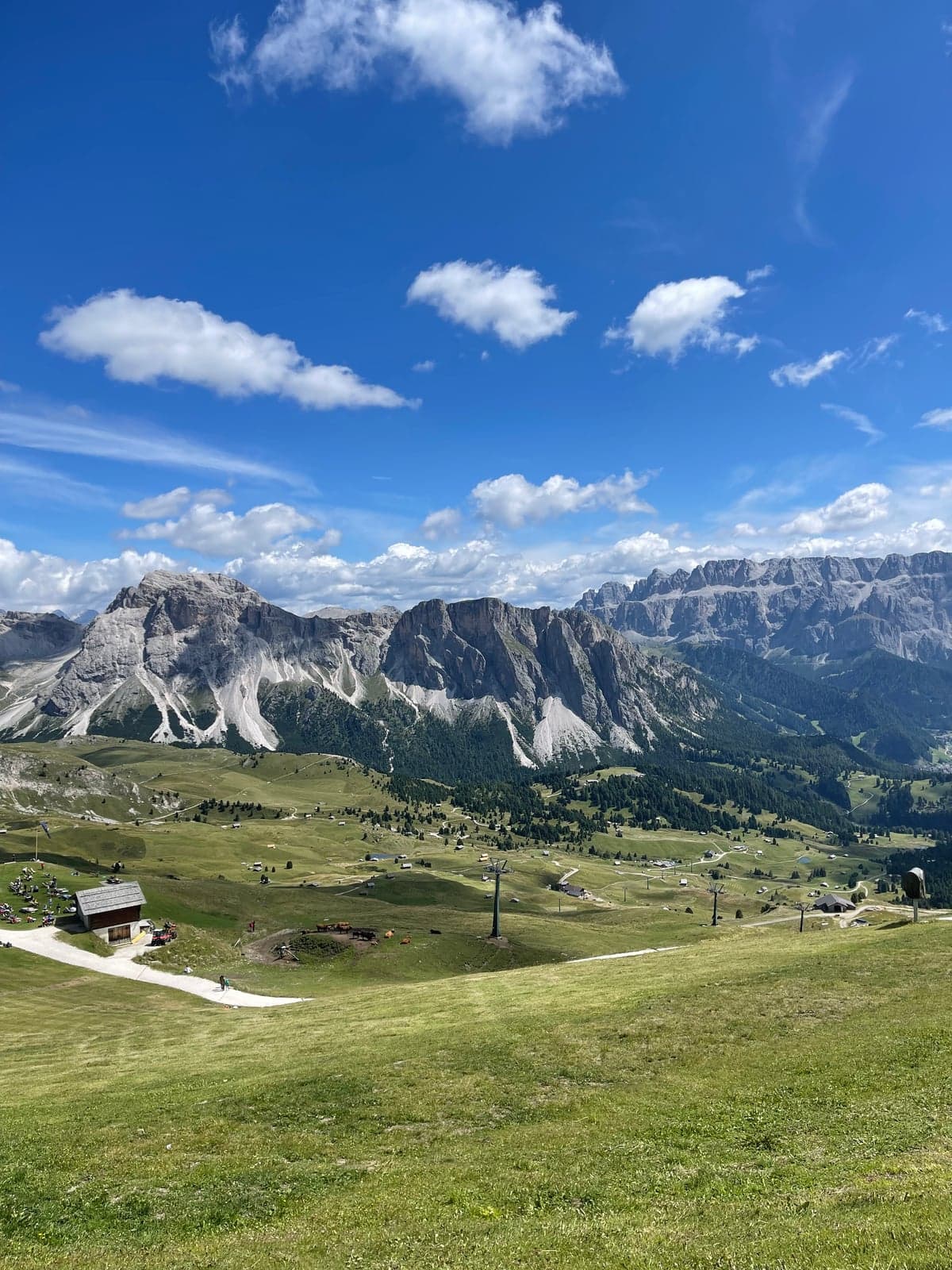 A meadow near the Seceda top station crossed by the poles of the Fermeda chairlift, hikers gathered by a hut, the Sella and Sassolungo groups rising along the horizon under a partly cloudy sky.