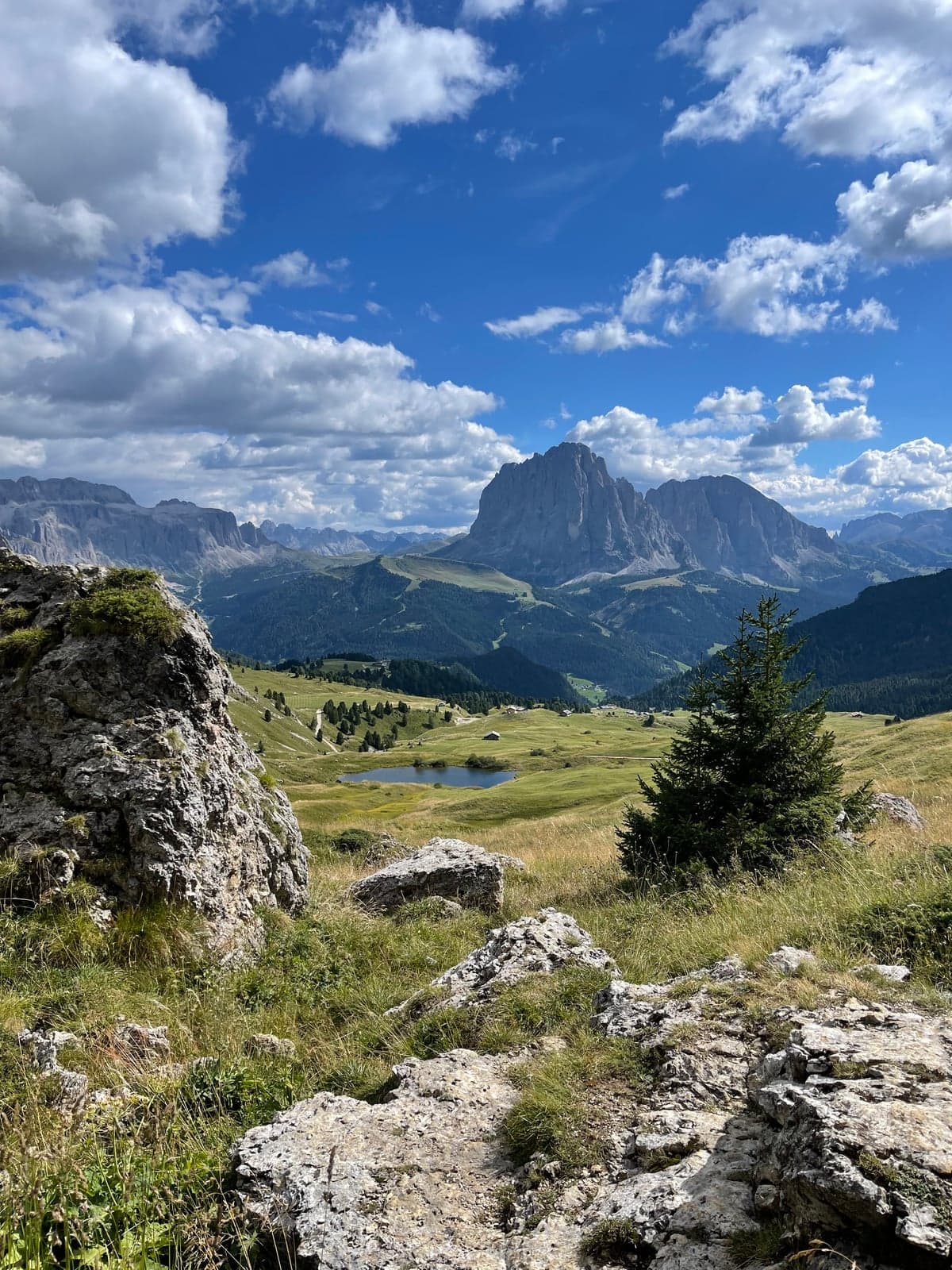 A small mountain tarn sits in an open meadow along a Seceda ridge footpath, with a jagged Dolomite peak rising on the horizon.