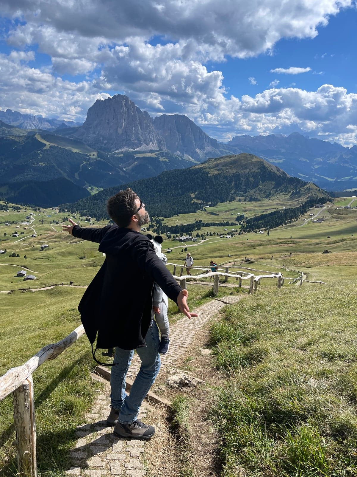 A hiker with a child on his back walks a paved footpath along the Seceda ridge, a weathered wooden fence running beside the trail and jagged Dolomite peaks filling the horizon.