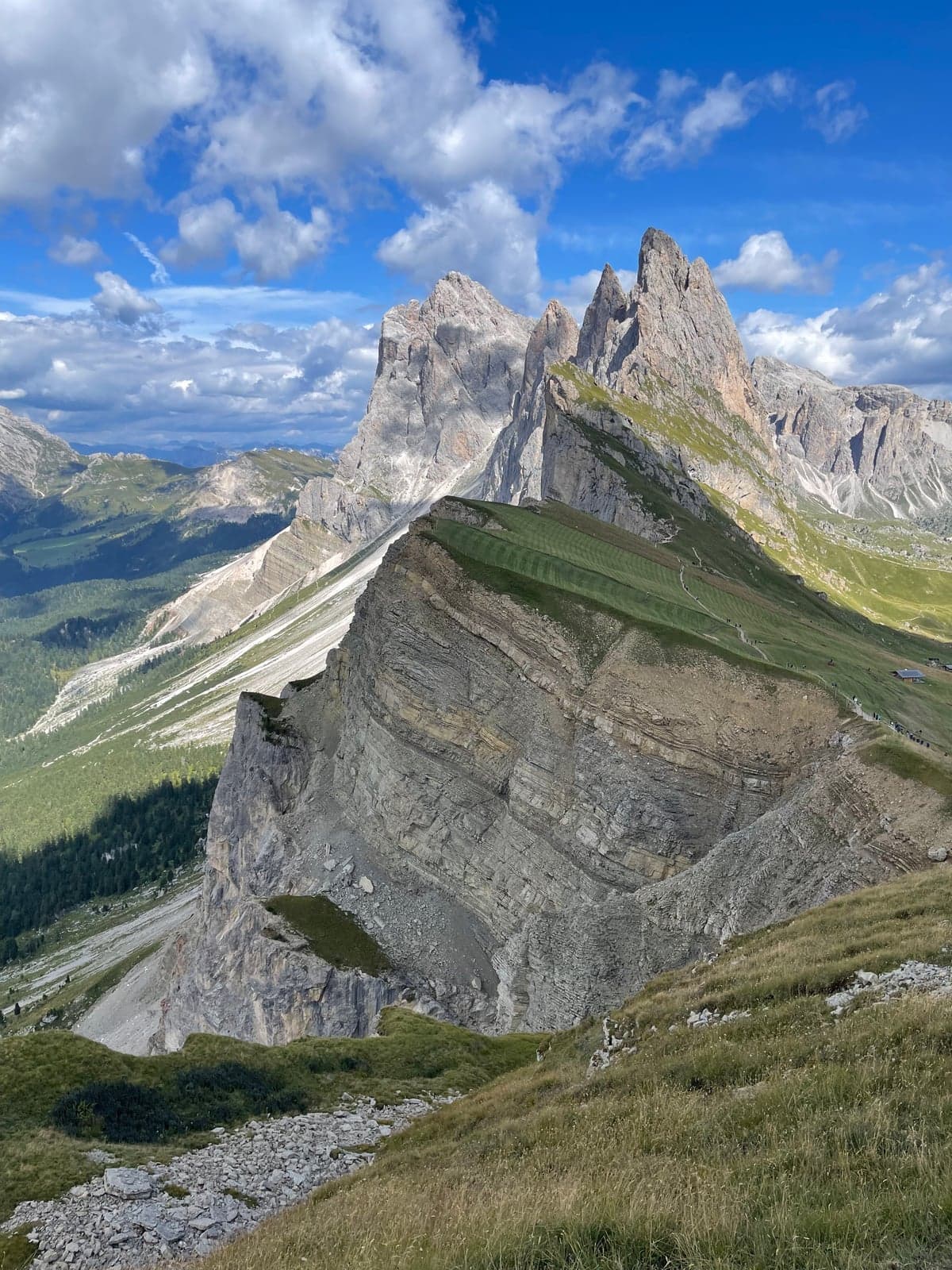 A grass ridge on Seceda curves away in a tilted blade toward the jagged Odle spires, tiny hikers visible along the ridge trail under a partly cloudy sky.