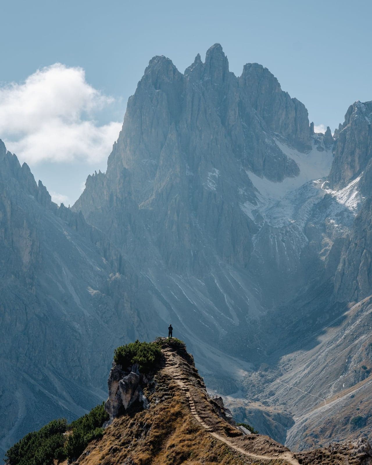 A lone hiker stands at the end of a narrow grassy spur at the Cadini di Misurina viewpoint, a worn footpath and rough steps running along its spine, with the jagged Cadini spires rising across the basin behind.