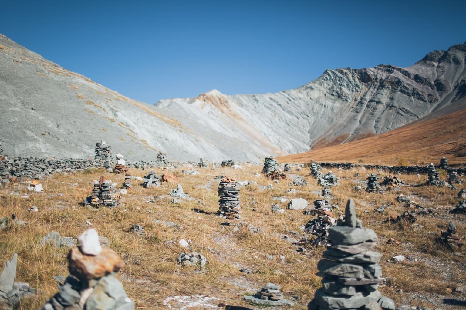 A dry alpine meadow scattered with dozens of small hand-stacked rock cairns, pale scree-covered mountainsides rising behind under a clear blue sky.