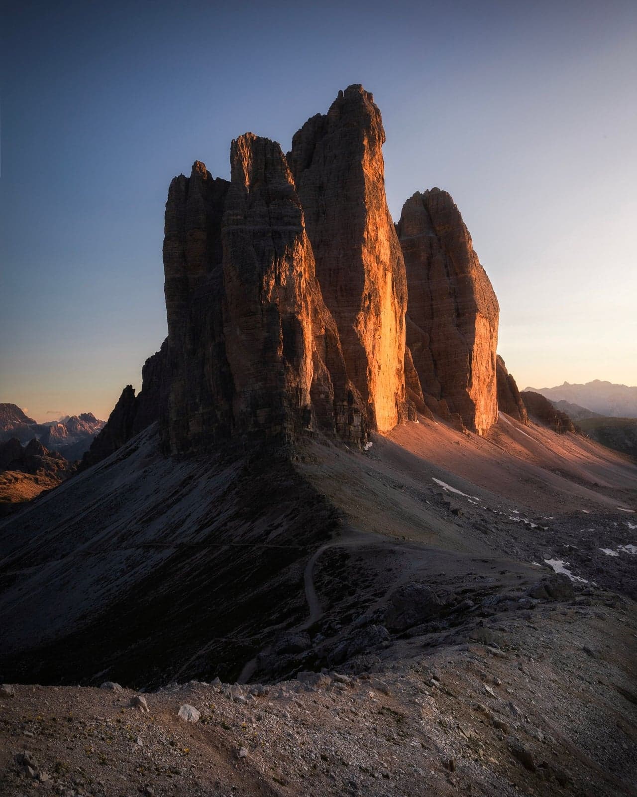 The three towers of Tre Cime di Lavaredo lit by warm, low-angle light, with an empty trail winding through the shadowed foreground, no people in sight.