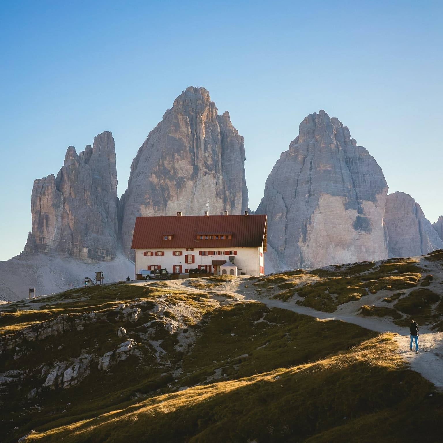 Rifugio Locatelli, a two-story alpine hut with a steep dark-red gabled roof, stands at the base of the three limestone towers of Tre Cime di Lavaredo, which rise directly behind it; a hiker walks the dirt trail beside the hut in late-day light.