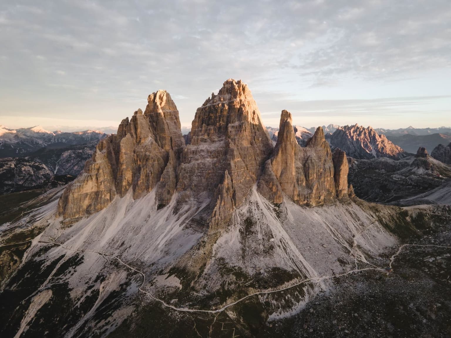 Aerial view of the Tre Cime di Lavaredo massif in warm, low-angle light, showing hiking trails circling the base of the three limestone towers from above.