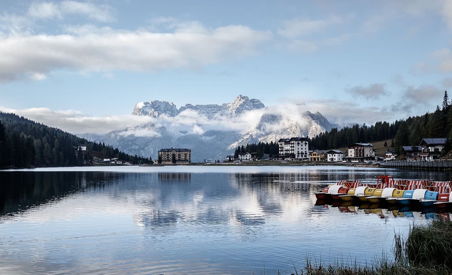 Lake Misurina under soft, low light, a row of colorful rental paddleboats moored at the shore, hotels lining the far bank, and snow-dusted Dolomite peaks emerging from cloud behind.