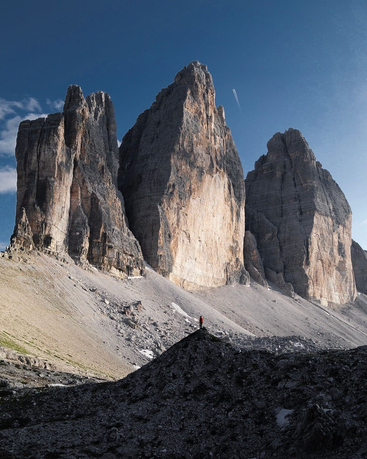 The three sheer limestone towers of Tre Cime di Lavaredo seen head-on in daylight under a blue sky, a lone hiker in a red jacket standing small on a rocky outcrop in the foreground for scale.