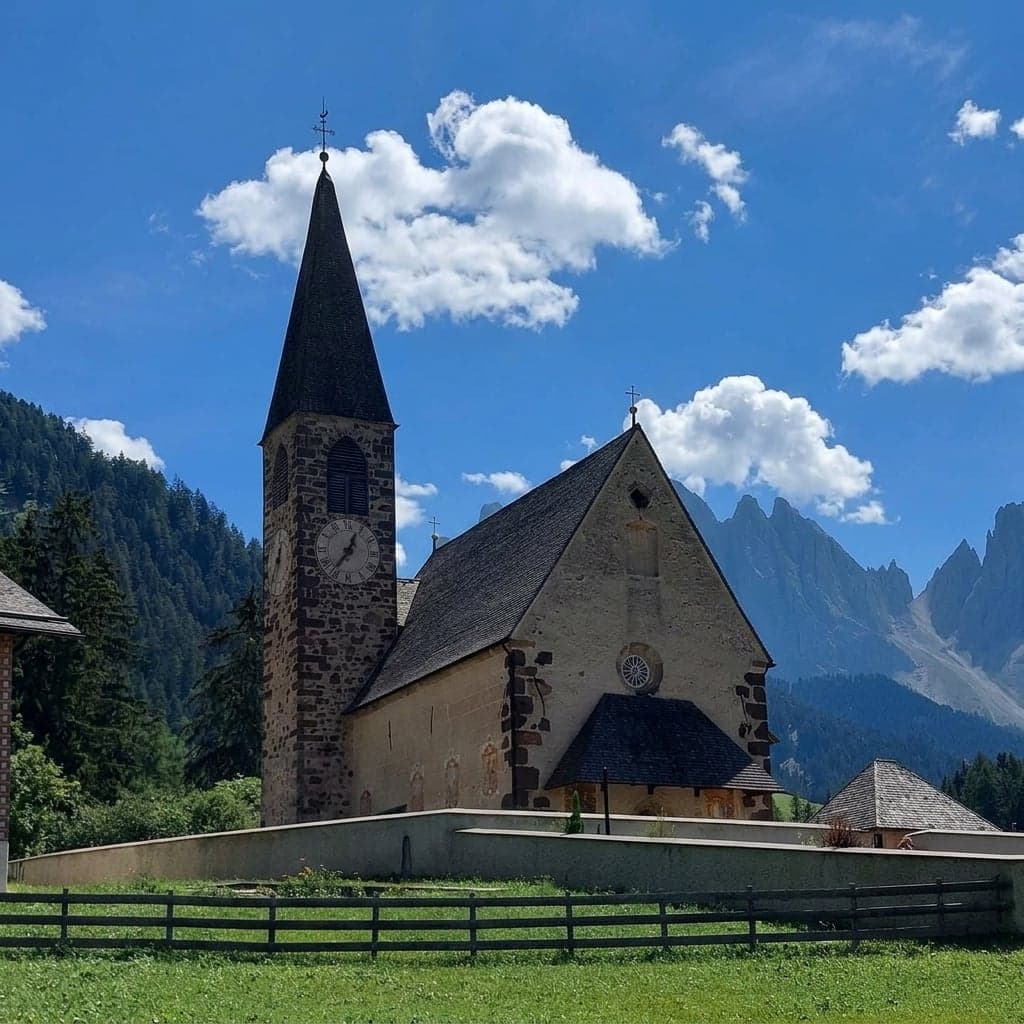 The church of Santa Maddalena filling the frame up close, its bell tower and clock visible, with the Odle peaks off to one side