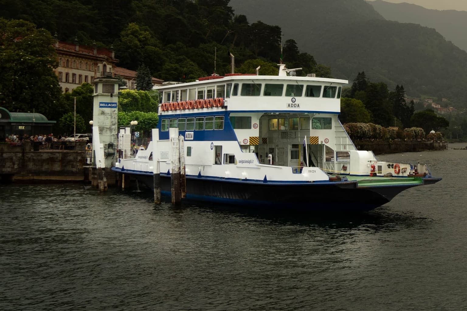 A Navigazione Laghi car ferry moored at the Bellagio ferry pier at dusk, with people on the promenade behind