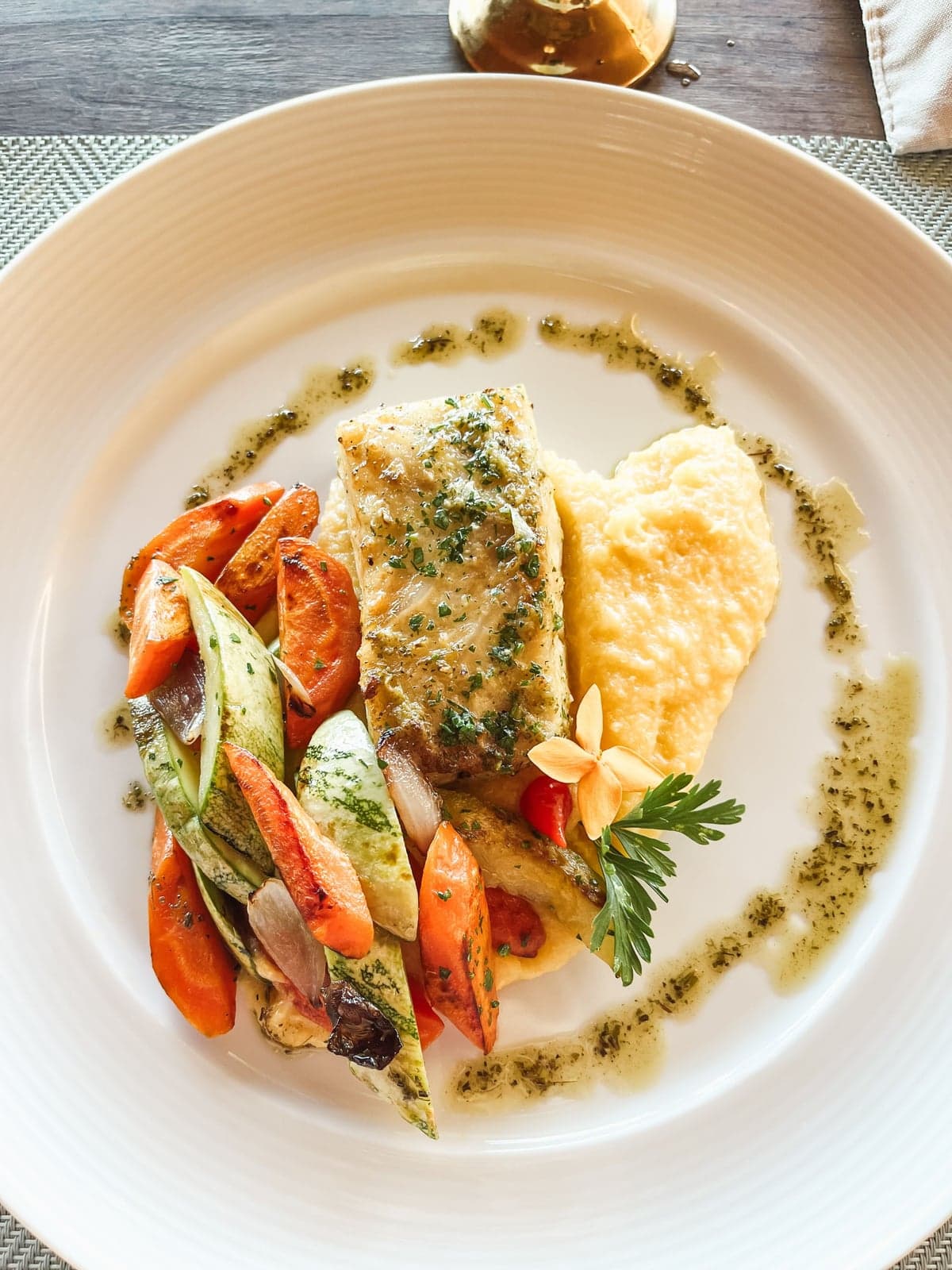 A fillet of lake fish with herb dressing, roasted vegetables and a scoop of soft polenta on a white plate