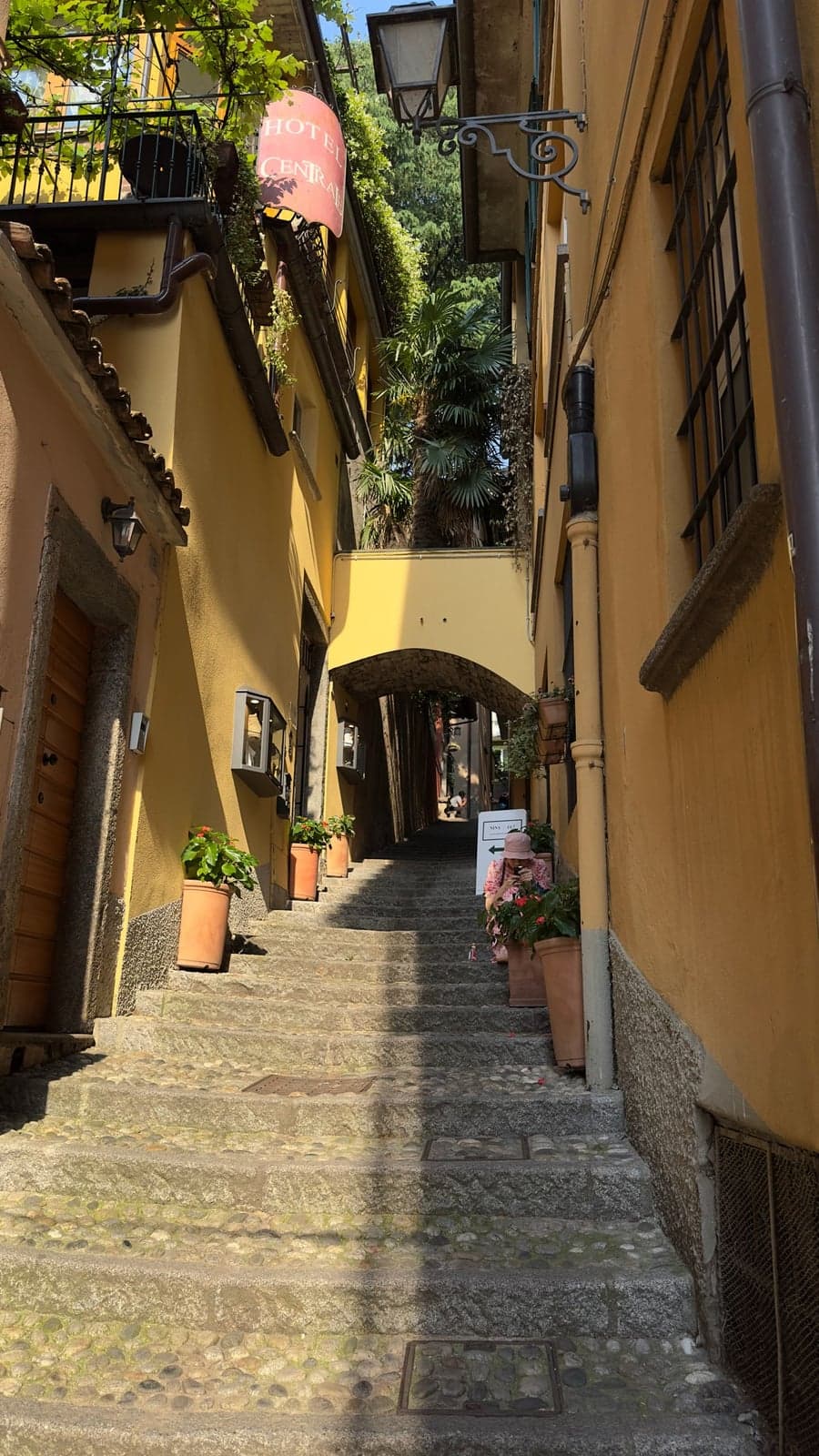 A quiet stepped back lane in Bellagio away from the crowds, potted geraniums and a small hotel sign along cobbled steps climbing between ochre houses