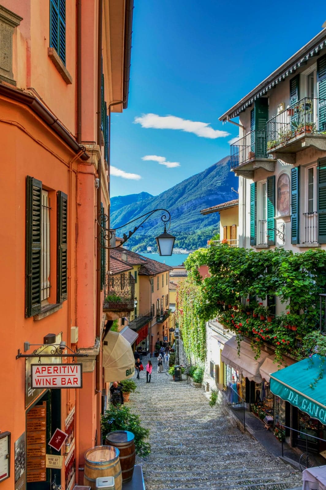 The stepped Salita Serbelloni lane in Bellagio, pastel buildings and cafe signs descending toward Lake Como with mountains beyond