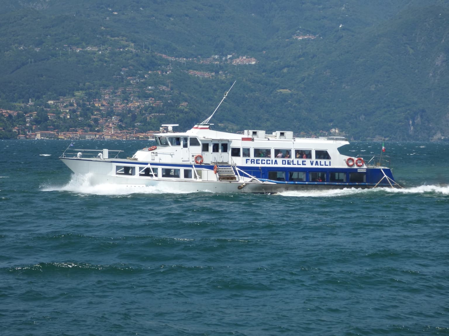 A white-and-blue Navigazione Laghi battello, the slow open-deck passenger ferry, crossing Lake Como