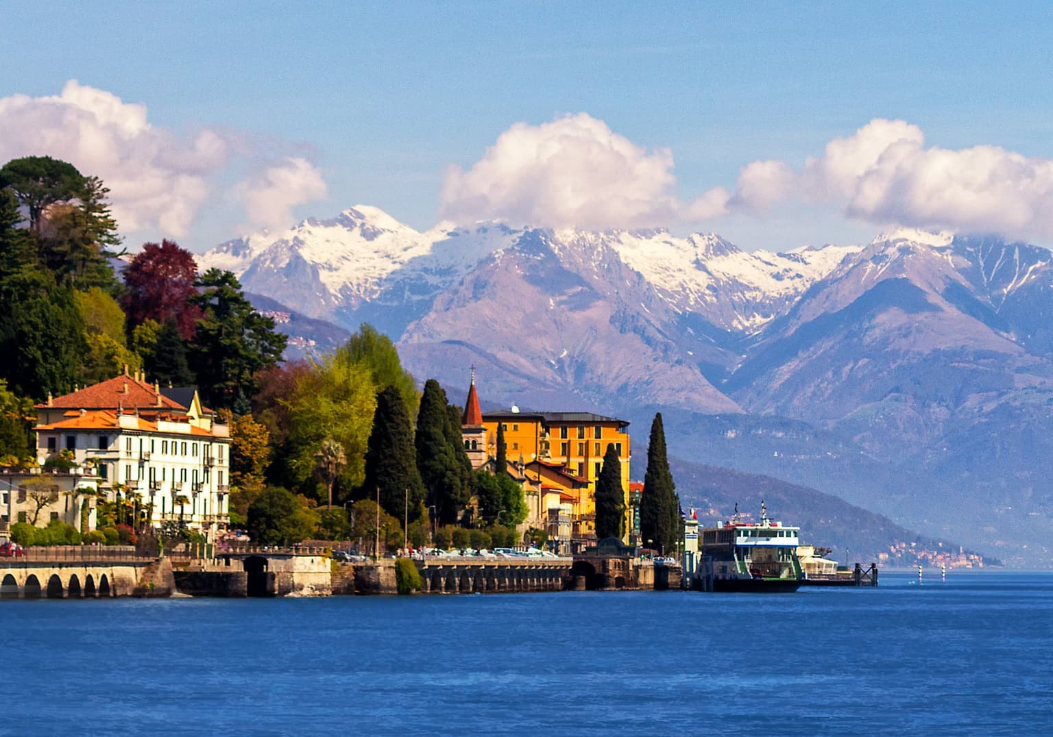 A Lake Como car ferry crossing the open water below snow-capped Alps, a lakeside villa on the western shore