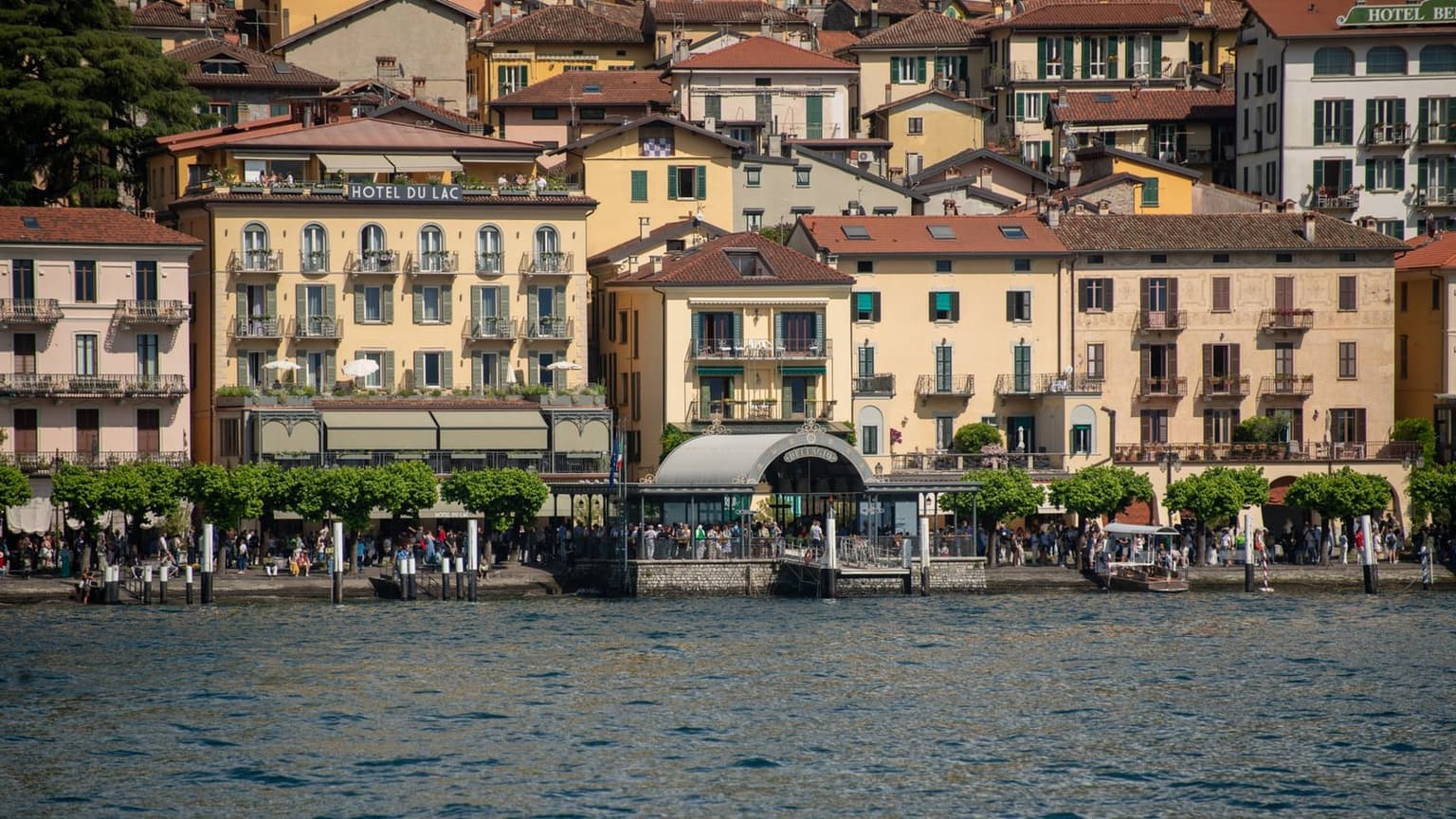 The Bellagio waterfront promenade packed with day-trippers at midday, crowds pressing along the lakefront beside the ferry dock