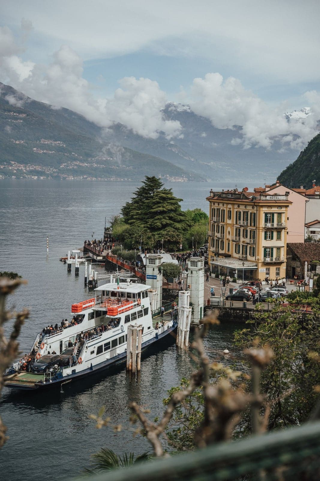 A Navigazione Laghi car ferry at the Varenna dock on Lake Como, vehicles on its deck and a crowd waiting on the pier below the yellow lakeside hotel