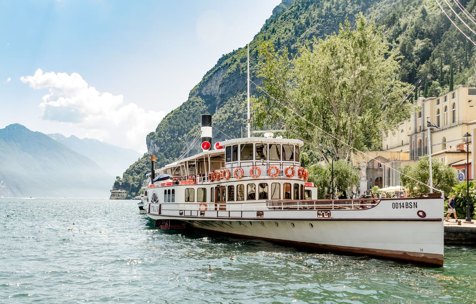 The historic paddle steamer Italia moored at a lakeside town pier on Lake Garda, a steep wooded cliff and an arcaded yellow building behind and mountains fading into haze across the water