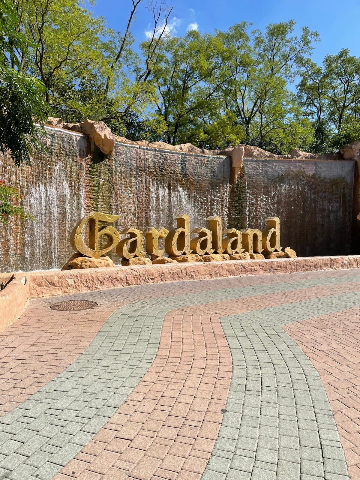 The Gardaland sign in large sculpted letters in front of a waterfall wall at the theme park entrance, trees above and a curving paved plaza in front