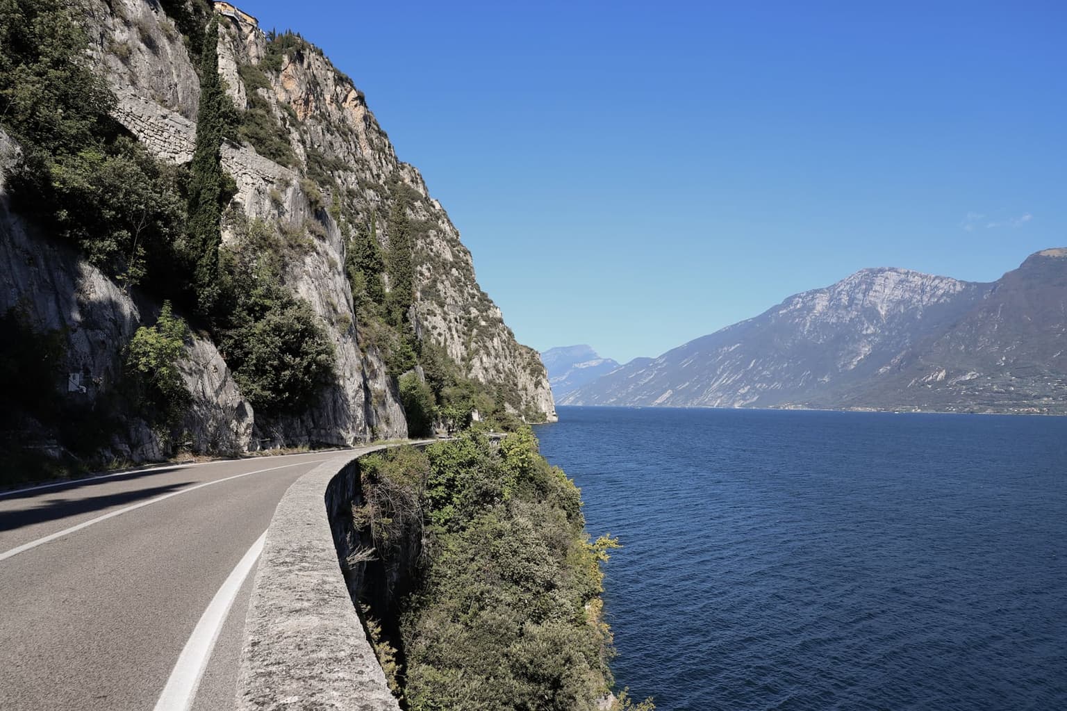 A narrow lakeside road cut into a sheer limestone cliff on Lake Garda, a stone parapet dropping straight to deep blue water and a high mountain ridge across the lake