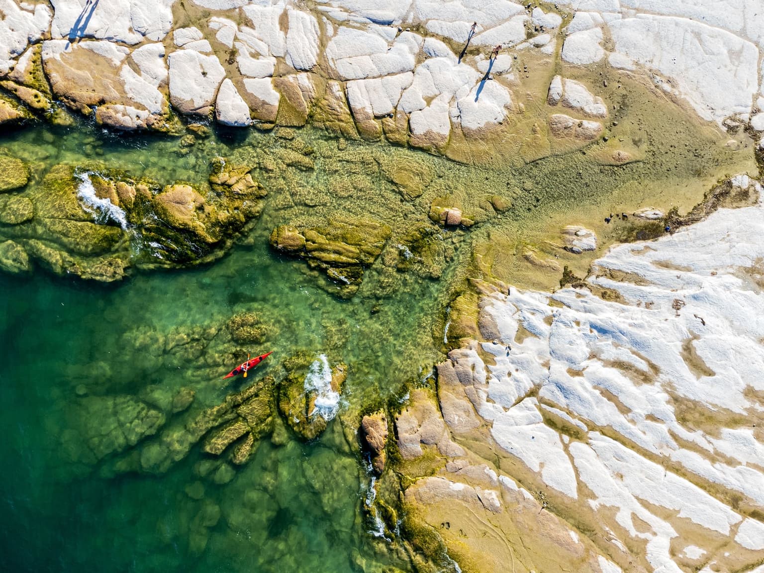 Looking straight down from the air at Jamaica Beach on the tip of the Sirmione peninsula, clear green shallows along a pale shore, a red kayak paddling past and a few people at the water's edge