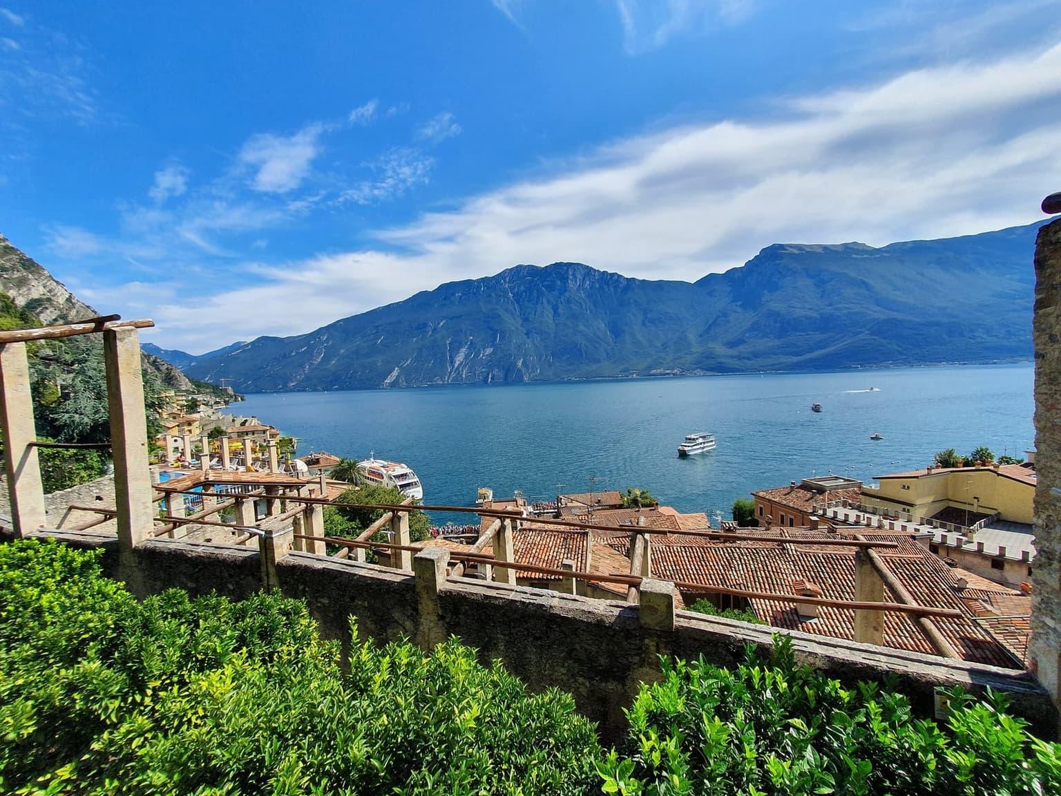Stone pillars and wooden beams of the Limonaia del Castel lemon house in Limone sul Garda, looking over terracotta rooftops to the lake, a white ferry near the harbor and the Monte Baldo ridge across the water