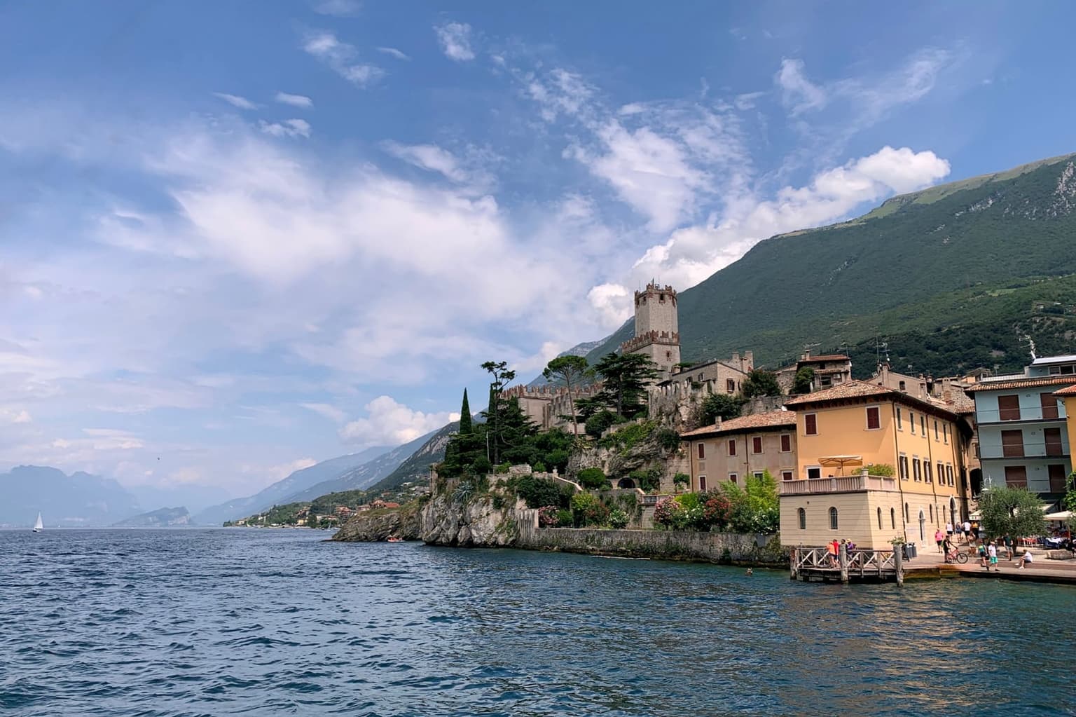 Malcesine seen from the lake, the Scaliger castle tower on a rocky spur above the water, ochre and yellow waterfront houses beside it and the forested slope of Monte Baldo rising behind