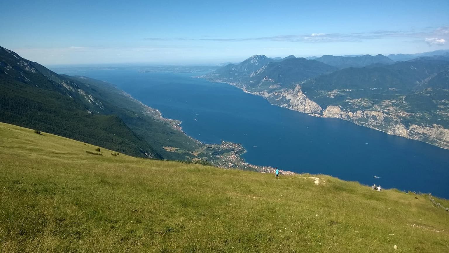 Lake Garda seen from a grassy ridge high on Monte Baldo, the deep blue lake stretching south between mountain shores, a lakeside town far below and two small hikers on the slope