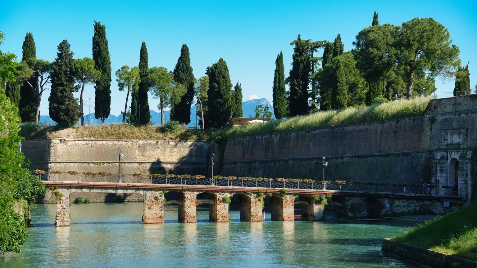 The Venetian walls of Peschiera del Garda rising from a turquoise canal, a low arched stone bridge in front and cypresses and pines along the top of the ramparts