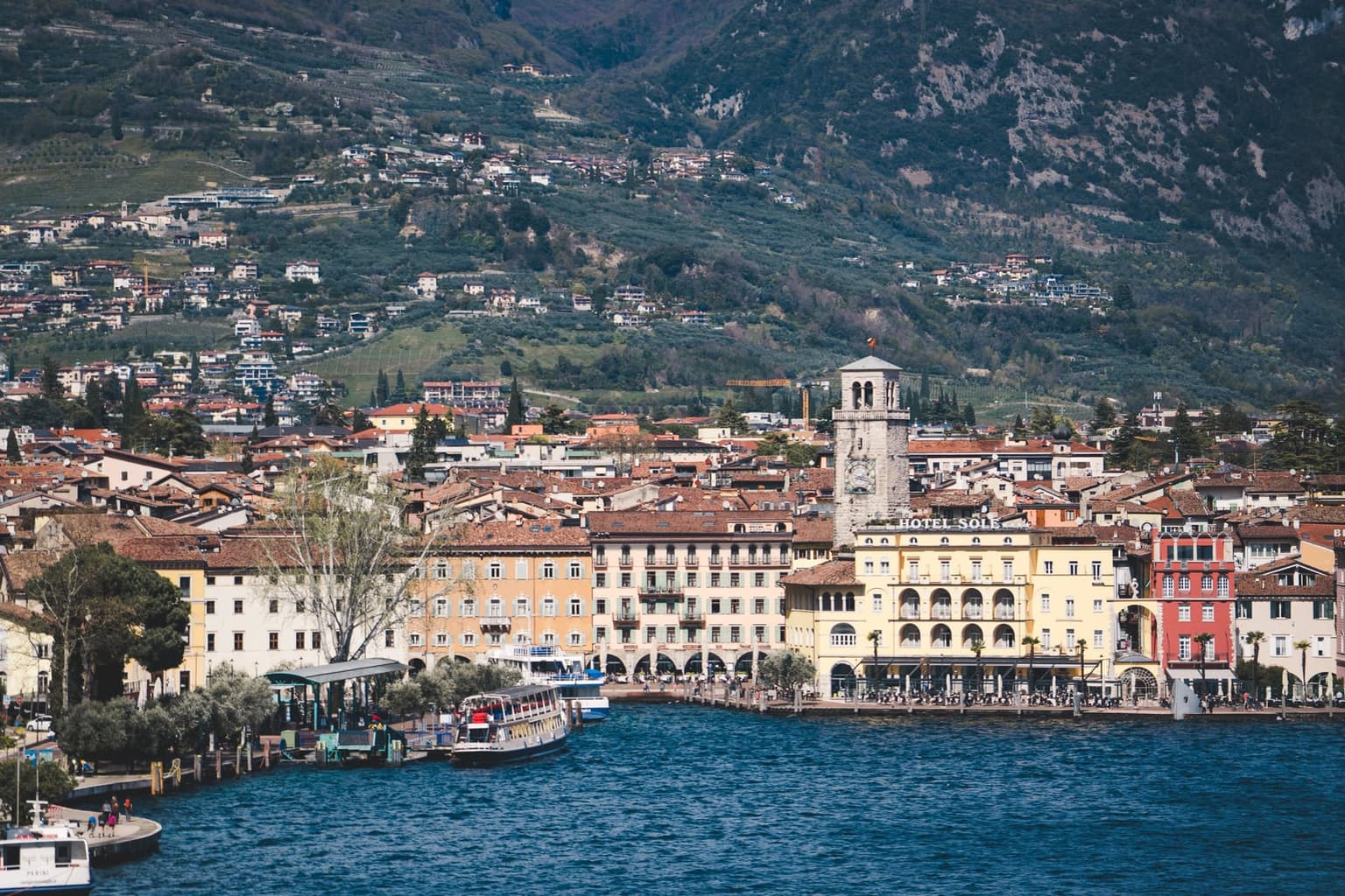 Riva del Garda's waterfront from the lake, the Torre Apponale clock tower above arcaded pastel buildings and the harbor boats, with houses climbing the steep mountainside behind