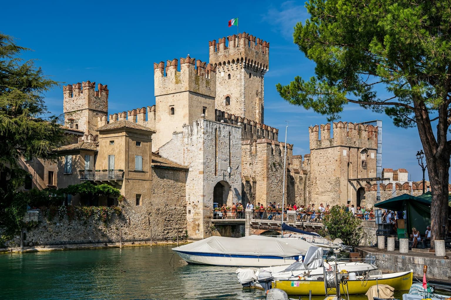 The Scaliger castle at Sirmione, crenellated stone towers rising above the moat, small boats moored in front and visitors crossing the entrance bridge under an Italian flag